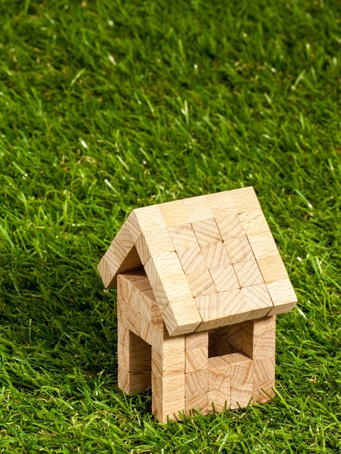 Eigentum: Ein Miniaturhaus, zusammengebaut aus kleinen Holzklötzen, steht in einer grünen Wiese. 